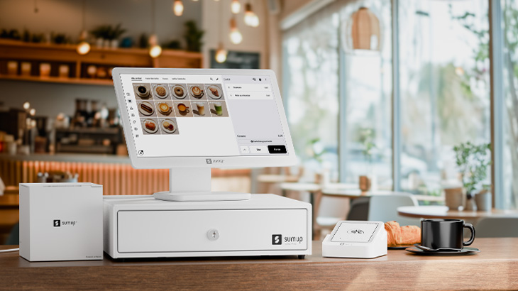 Modernes weißes SumUp-Kassensystem mit Touchscreen und Kartenleser auf einer Theke in einem Café, im Hintergrund unscharf Tische, Lampen und große Fenster mit Tageslicht.