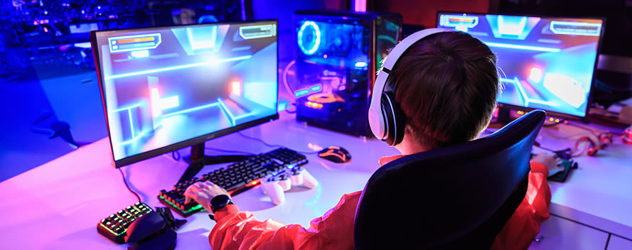 Ein Junge mit Gaming Headset zockt ein PC Spiel an zwei Bildschirmen und bedient dabei die Tastatur während der Controller daneben liegt.