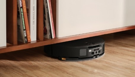 Schwarzer Roboterstaubsauger unter einem Holzregal auf einem Holzboden, neben Büchern.