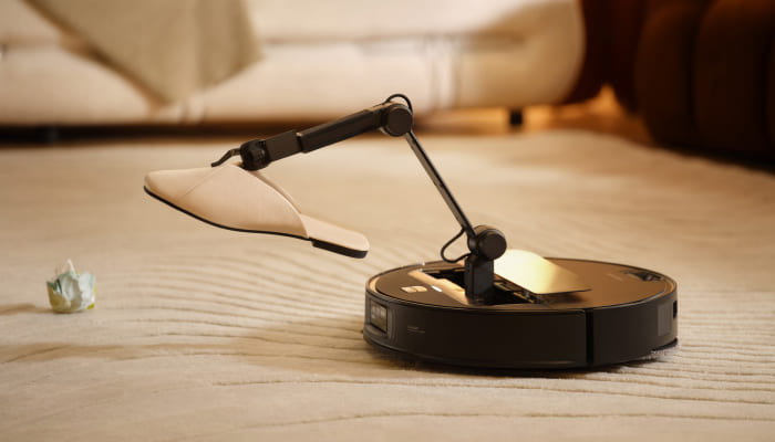 Ein Roboterstaubsauger mit einem montierten Roboterarm hält einen Schuh über einem Teppich, neben einem zerknitterten Papier.