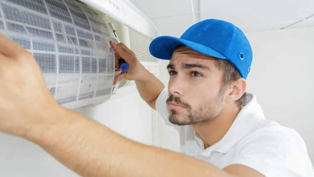 Ein Mann mit blauer Kappe wartet eine Klimaanlage mit einem weißen Filter in einem weißen Raum.