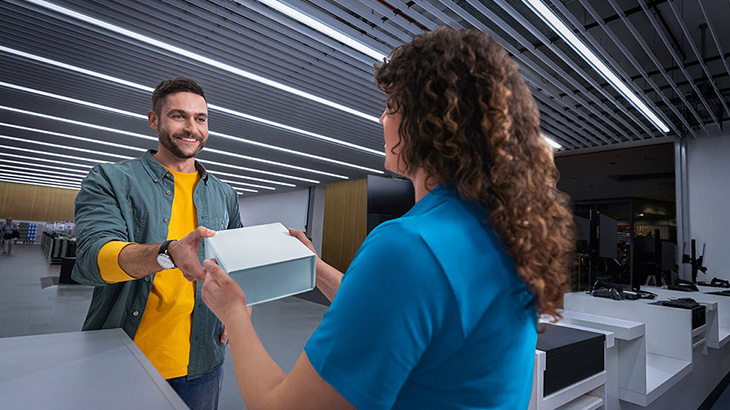 Ein lächelnder Mann in gelbem Hemd erhält von einer Person in Blau eine weiße Schachtel an einem Tresen.