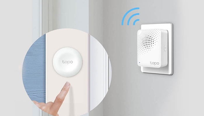 Ein Finger drückt einen runden, weißen "Topo"-Knopf an einer Wand neben einem Türrahmen. Rechts ein weißer "Topo"-Stecker mit blauen WLAN-Symbolen.