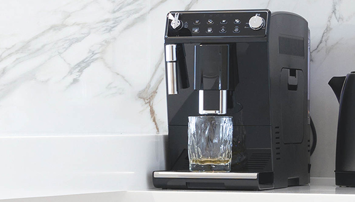 Eine schwarze Kaffeemaschine steht auf einer weißen Arbeitsfläche vor einer Marmorwand, ein Glas darunter.