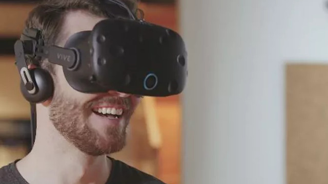 Mann lächelt mit VR-Headset und Kopfhörern, trägt ein graues T-Shirt vor einem neutralen Hintergrund.