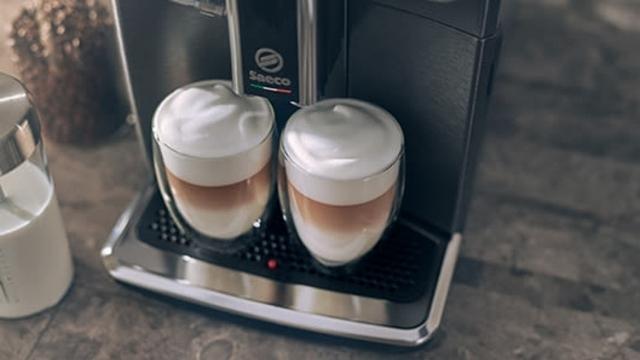 Zwei Gläser Latte Macchiato mit Milchschaum in einer Kaffeemaschine. Ein Milchbehälter steht daneben auf einer grauen Oberfläche.