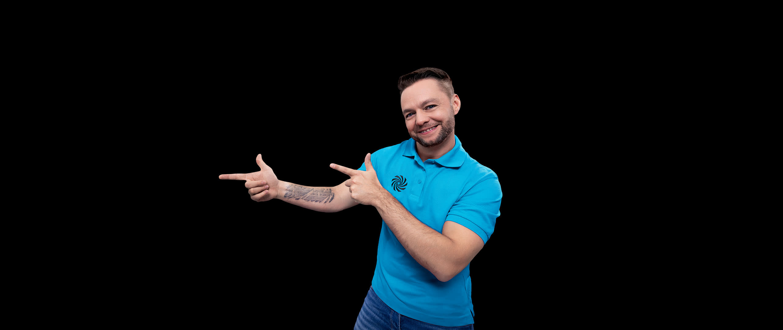 Ein Mann in blauem Polohemd und Jeans lächelt und zeigt mit beiden Händen auf etwas, im Hintergrund ist Schwarz.