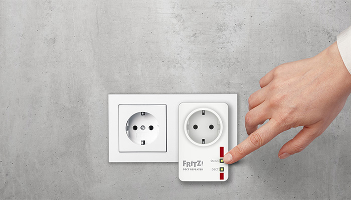 Eine Hand drückt einen Knopf an einem weißen FRITZ! WLAN Repeater, der in einer Steckdose an einer grauen Wand steckt.