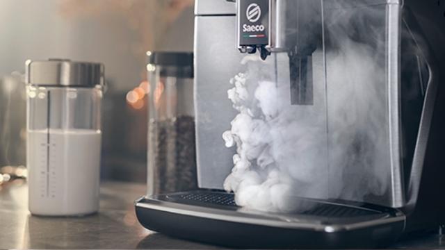 Eine Saeco Kaffeemaschine gibt Dampf ab, neben Behältern mit Milch und Kaffeebohnen auf einer Arbeitsfläche.