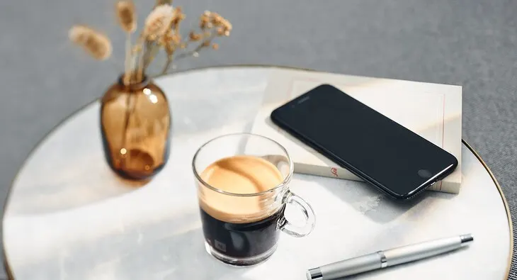 Auf einem Tisch sind eine braune Vase mit Pflanzen, ein Kaffee im Glas, ein schwarzes Handy auf einem Buch und ein Stift zu sehen.