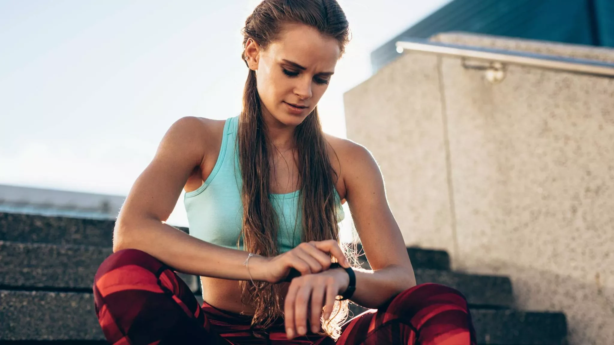 Eine Sportlerin sitzt auf einer Treppe draußen und tippt auf ihre schwarze Smartwatch.