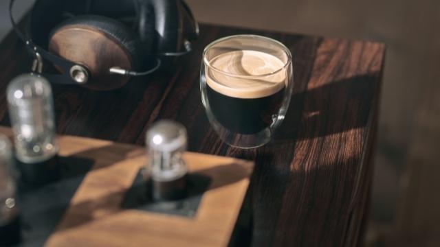 Auf einem Holztisch steht ein Glas mit dunklem Kaffee und hellem Schaum, neben Kopfhörern und Verstärkern.