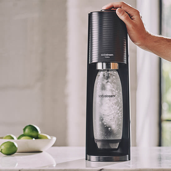 Eine Hand bedient einen SodaStream Wassersprudler mit einer Flasche. Eine Schale mit Limetten steht daneben.