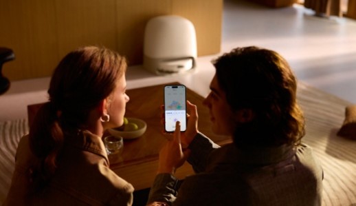 Zwei Personen sitzen an einem Tisch und schauen auf ein Smartphone. Im Hintergrund steht ein weißes Gerät.