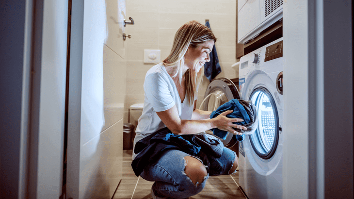 Frau in Jeans und T-Shirt hockt vor Waschmaschine und hält blaue Wäsche. Im Hintergrund sind Toilette und Handtücher zu sehen.