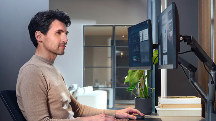 Ein Mann sitzt an einem Schreibtisch mit zwei Computermonitoren, einer Pflanze und Büchern.