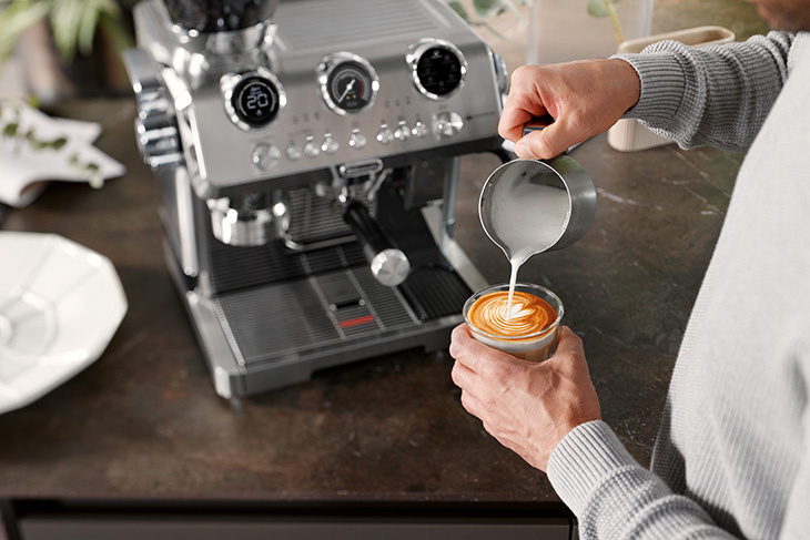 Person gießt feinporigen Milchschaum in einen Latte Art Kaffee neben einer modernen Siebträgermaschine.