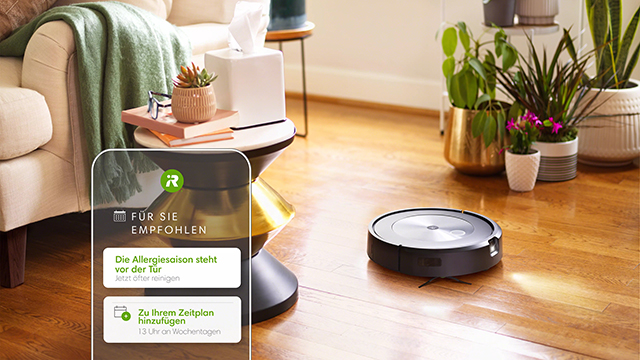 Ein runder Staubsaugerroboter auf einem Holzboden, ein Sofa mit grüner Decke, Pflanzen und ein Smartphone.