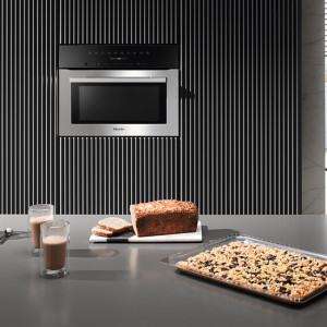 Einbauofen an gestreifter Wand über Tisch mit Brot, Kuchen und Getränken in Gläsern auf einer grauen Oberfläche.