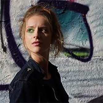 Eine Person in dunkler Jacke steht vor einer Wand mit Graffiti in Schwarz, Weiß und Blau.