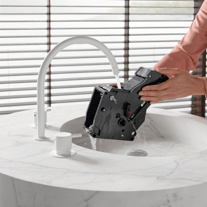 Eine Person reinigt eine schwarze Kaffeemaschinenkomponente unter einem Wasserhahn in einem weißen Waschbecken. Jalousien im Hintergrund.