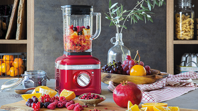 Ein roter Mixer gefüllt mit Früchten steht auf einer Theke, umgeben von frischen Früchten und Küchenutensilien vor Regalen.