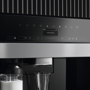 Nahaufnahme einer schwarzen Kaffeemaschine mit einem Glas Milch und einem leeren Glas darunter. Silberne Akzente sind sichtbar.