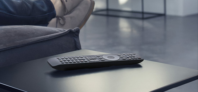 Eine schwarze Fernbedienung liegt auf einem dunklen Tisch, im Hintergrund ist ein Teil eines Sofas zu sehen.