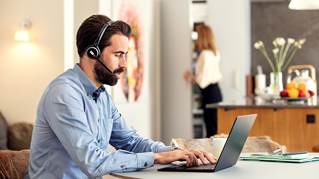 Mann mit Headset tippt auf Laptop. Frau im Hintergrund, Tisch mit Blumen und Obst.