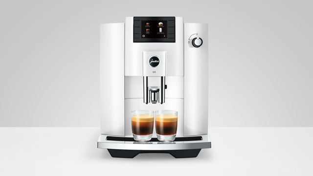 Ein weißer Kaffeevollautomat mit zwei Gläsern braunem Kaffee steht auf einer weißen Oberfläche vor einem hellgrauen Hintergrund.