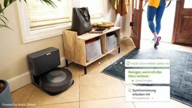 Ein Raum mit einem Roboterstaubsauger neben einer Kommode, auf der eine Tasche liegt. Eine Person geht durch eine offene Tür.