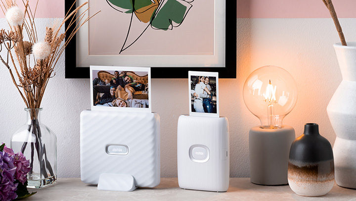 Zwei weiße Drucker drucken Fotos. Es gibt eine Lampe, zwei Vasen und ein gerahmtes Bild auf einem Tisch vor einer Wand.