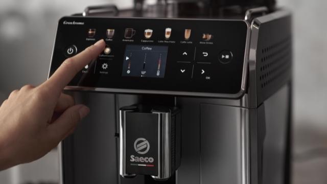Eine Hand berührt das Display einer Saeco Kaffeemaschine. Das Display zeigt Kaffeeoptionen und Einstellungen.