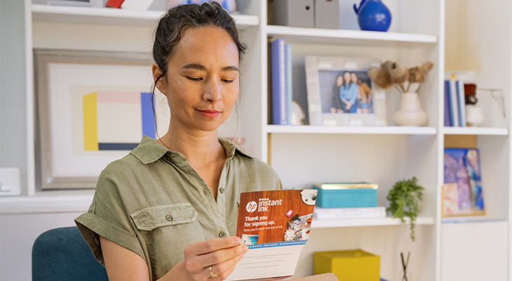 Eine Frau in einem grünen Hemd hält eine HP Instant Ink Karte vor einem weißen Bücherregal.