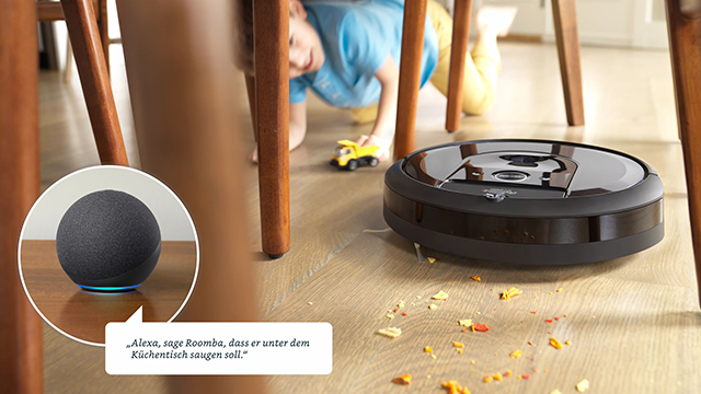 Ein schwarzer Saugroboter reinigt einen Holzboden. Ein Kind spielt mit Spielzeug unter einem Tisch. Ein Echo-Gerät steht daneben.