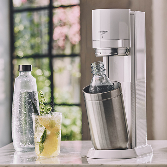 Ein Sodastream-Gerät steht neben einer Flasche Wasser und einem Glas mit einem Getränk auf einem Tisch.