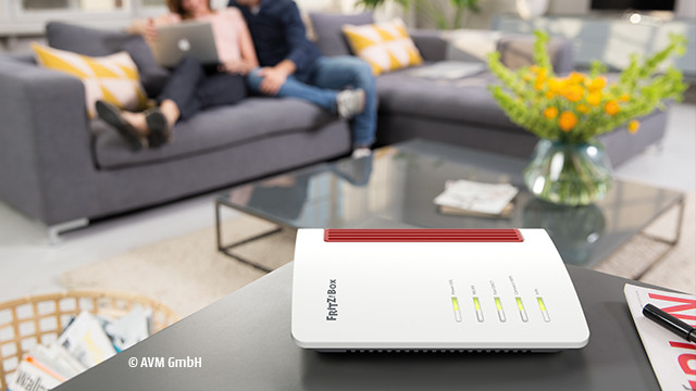 Ein weißer Router mit roten Akzenten steht auf einem Tisch, im Hintergrund sitzt ein Paar auf einem grauen Sofa.