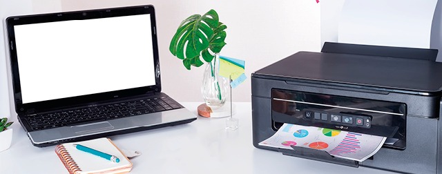 Ein Schreibtisch mit einem Laptop mit leerem Bildschirm, einem Drucker, einer Pflanze in einer Vase, einem Notizblock und einem Stift.