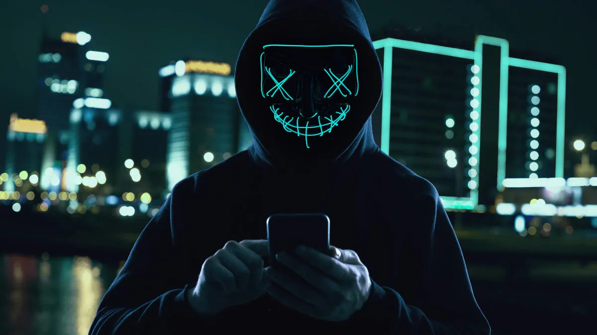 Eine Person mit einer leuchtenden Maske und Kapuzenpullover hält ein Smartphone vor einer Stadtkulisse bei Nacht.