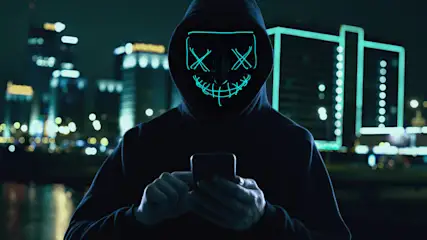 Eine Person mit einer leuchtenden Maske und Kapuzenpullover hält ein Smartphone vor einer Stadtkulisse bei Nacht.