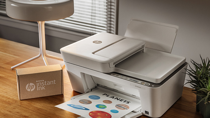 Ein Drucker druckt ein Blatt mit bunten Planeten. Daneben eine Lampe, eine Tintenbox und eine Topfpflanze auf einem Holztisch.