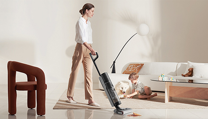 Eine Frau saugt Staub mit einem Staubsauger, während ein Kind und ein Hund im Hintergrund sitzen. Ein brauner Stuhl ist auch sichtbar.
