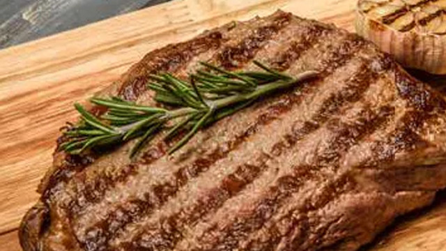 Ein Steak mit Grillstreifen und einem Rosmarinzweig liegt auf einem Holzbrett, daneben eine Knoblauchknolle.