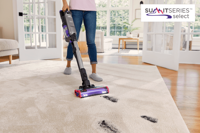 Eine Person saugt mit einem Staubsauger einen hellen Teppich. Im Hintergrund sind Möbel und ein Fenster zu sehen.