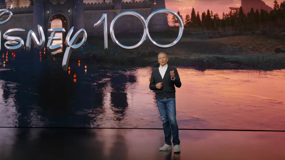 Ein Mann in Jeans und Strickjacke gestikuliert vor einem Hintergrund mit Schloss, Fluss und dem Schriftzug "Disney 100".