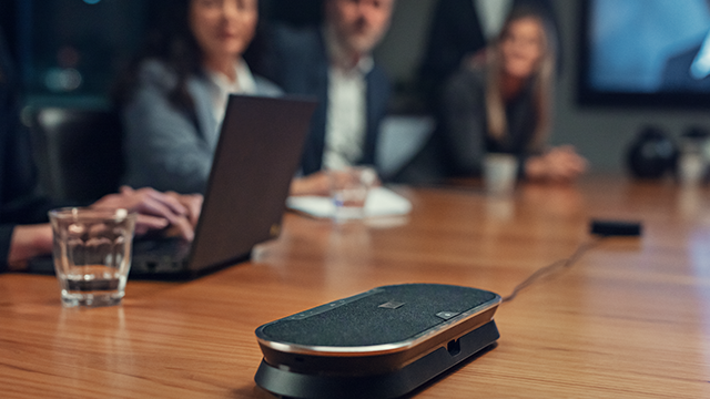 Ein Konferenztisch mit einem Konferenztelefon, einem Laptop, einem Glas Wasser und mehreren Personen im Hintergrund.