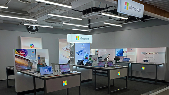 Ein Innenraum zeigt mehrere Tische mit Laptops unter der Beleuchtung, mit Microsoft-Logos und Werbedisplays.