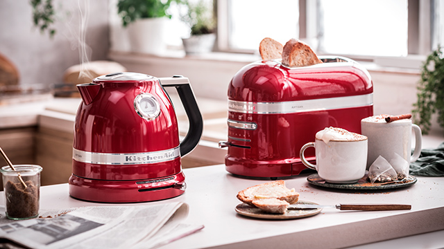 Ein roter Wasserkocher und Toaster stehen auf einer Theke neben Tassen, Toast und einer Zeitung.
