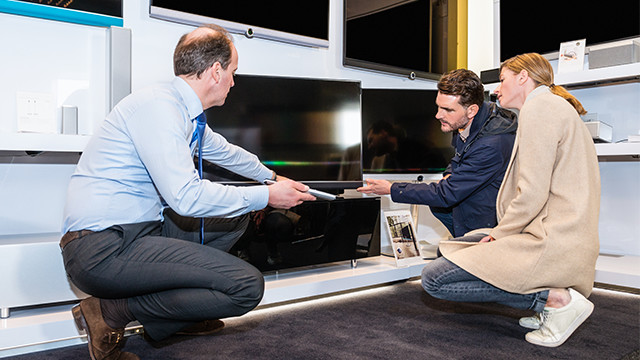 In einem Laden hockt ein Paar vor einem Fernseher, den ein Verkäufer mit einer Fernbedienung bedient.