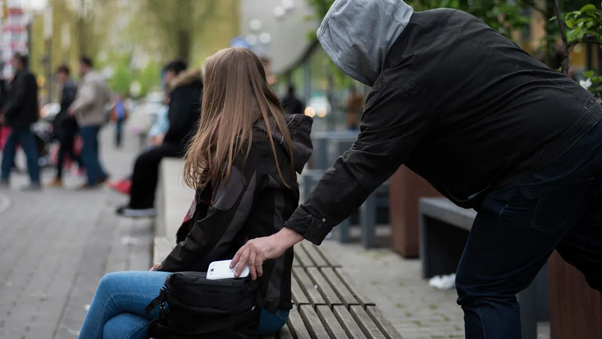 Eine Frau sitzt auf einer Bank, während eine Person ihr Handy aus der Tasche stiehlt. Passanten sind im Hintergrund zu sehen.